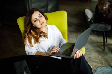 Genç, sempatik bir kadın, ince başlı bir vücut bina değil, rahat bir kafede oturur ve bir dizüstü bilgisayar için çalışır. İş giyim tarzı.