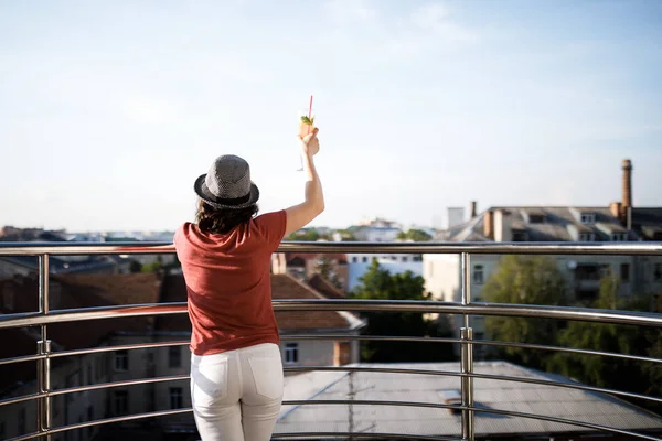 La jeune jolie brune se tient sur le balcon et tient une boisson fraîche dans ses mains. Magnifique vue sur la ville — Photo