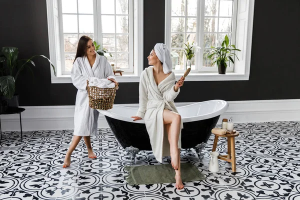 可爱的年轻两个女人穿着白色睡衣和毛巾在头上有乐趣在浴室 — 图库照片