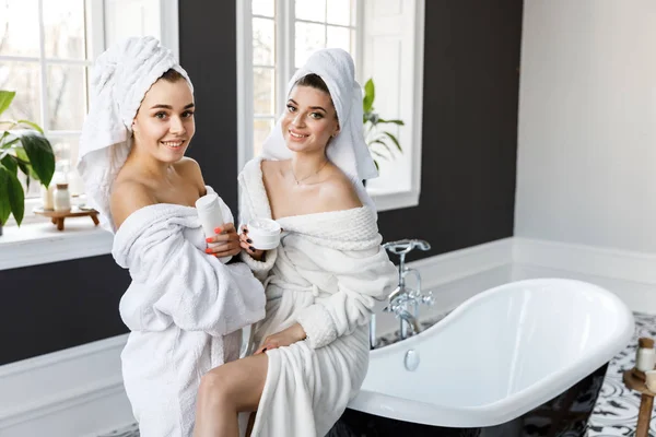 Niedliche junge zwei Frauen in weißen Morgenmänteln und Handtüchern auf dem Kopf haben Spaß im Badezimmer — Stockfoto