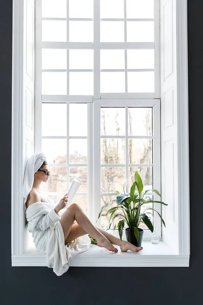 A cute young woman dressed in a white robe and with towels on her head sitting on the window sill and reading a magazine