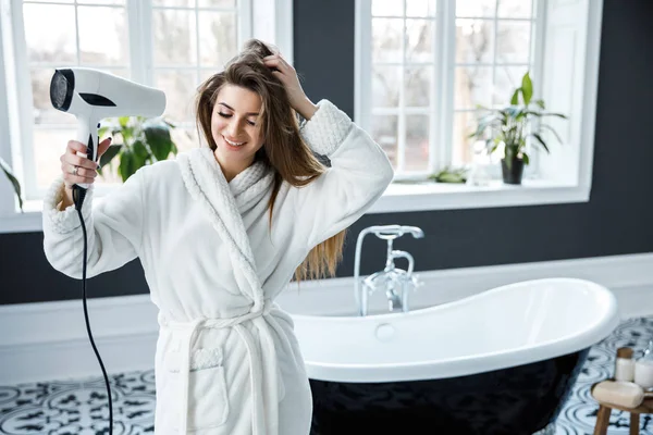 Young pretty woman dressed in white dressing gown drives her hair with a hair dryer in a beautiful bathroom.