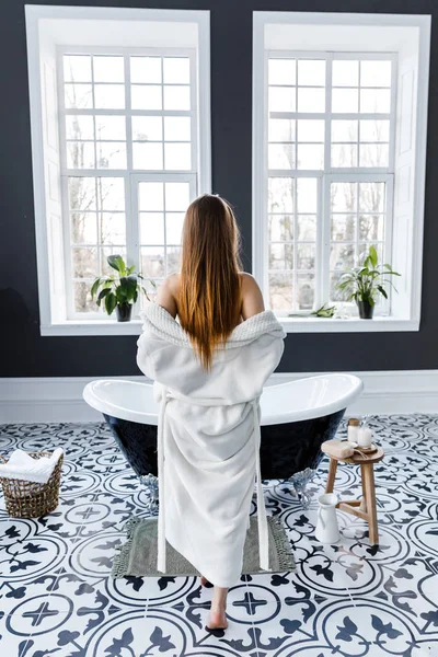 美丽的浅色浴室,有大窗户。一位年轻女子站在浴缸边脱下她的白色长袍 — 图库照片