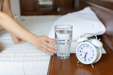 Ahşap bir başucunda beyaz bir çalar saat ve bir bardak su bulunmaktadır. Sabah. Bir kadın elini bir bardak suya uzatır.