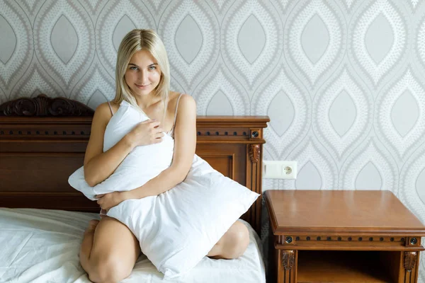 Positive Frau mit Kopfkissen sitzt im Bett und lächelt zu Hause in die Kamera. froher Morgen. — Stockfoto