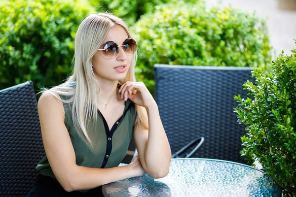 Jeune belle blonde assise dans un café au milieu de la ville en lunettes. La fille est bien habillée, style business. La fille est prête pour une réunion d'affaires. — Photo