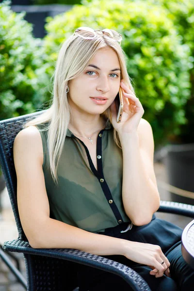 En ung vacker blond flicka sitter i ett kafé mitt i staden och pratar över telefon. Flickan är välklädd, Business Style. — Stockfoto