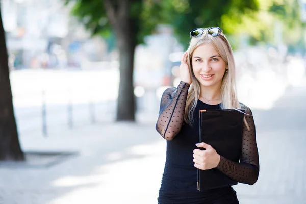 En ung vacker blond flicka klädd i en klassisk svart klänning innehar en mapp med papper i hennes händer. Affärskläder stil. — Stockfoto