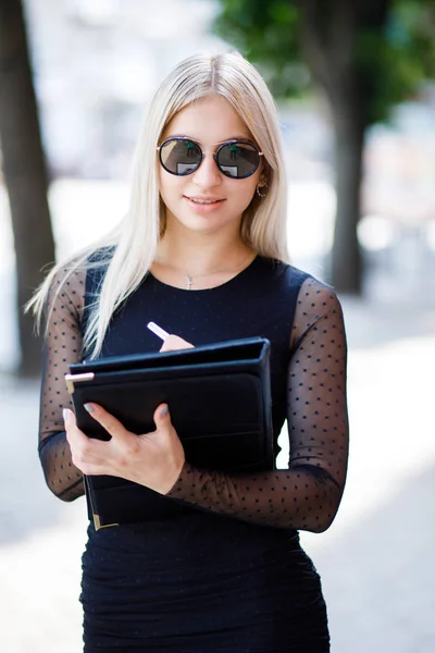 Uma jovem linda garota loira vestindo um vestido preto clássico tem uma pasta de papéis em suas mãos. Estilo de vestuário de negócios . — Fotografia de Stock