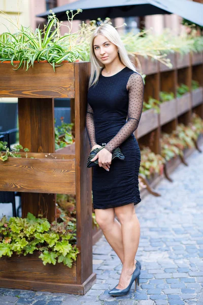 La jeune belle blonde, vêtue d'une robe noire classique, tient dans ses mains un petit sac à main, se promène dans les rues de la ville. Style de vêtements d'affaires — Photo