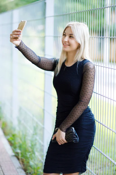 La jeune belle blonde, vêtue d'une robe noire classique, tient dans ses mains un petit sac à main et un smartphone, se promène dans les rues de la ville. Style de vêtements d'affaires . — Photo