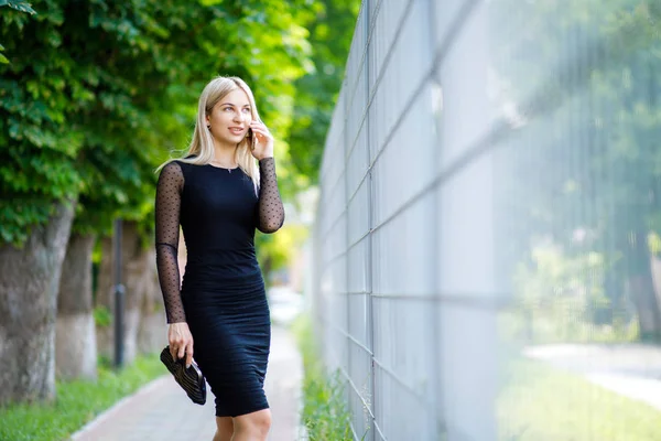 La joven y hermosa rubia, vestida con un vestido negro clásico, sostiene un pequeño bolso y un teléfono inteligente en sus manos, camina por las calles de la ciudad. Estilo de ropa de negocios . —  Fotos de Stock