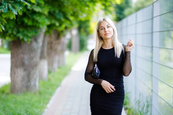 Den unga vackra blonda flickan, klädd i en klassisk svart klänning, rymmer en liten handväska i händerna, går genom gatorna i staden. Affärskläder stil — Stockfoto