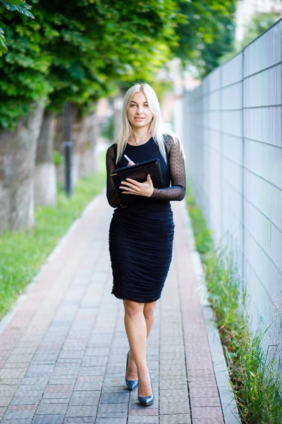 Uma jovem linda garota loira vestindo um vestido preto clássico tem uma pasta de papéis em suas mãos. Estilo de vestuário de negócios . — Fotografia de Stock