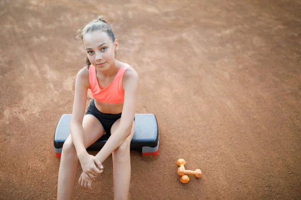 A cute, pretty teenage girl sits on a step platform and relaxes after her workout on outdoor — Stock Photo, Image