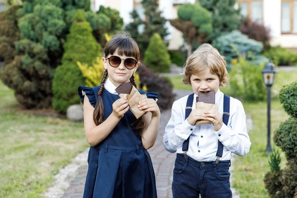 Cute blond guy and cute girl, school year fun time outdoors. Children eat chocolate. Children dressed in school uniform play games in the park