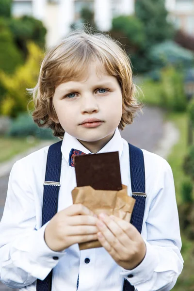 Cute blond guy, school years dressed in school uniform with pleasure eating black chocolate on the street in the park