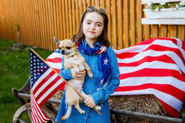 The girl stands in the yard, dressed in a denim dress and holding the flag of USA and little cute dog clipart