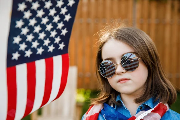 Дівчинка стоїть на подвір "ї, одягнена в джинсову сукню і тримає прапор США. — стокове фото