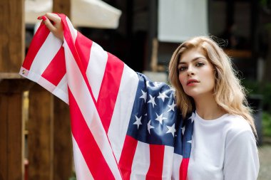 Genç ve güzel bir kız sokakta duruyor ve elinde büyük bir Amerikan bayrağı tutuyor.