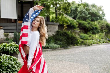 Genç ve güzel bir kız sokakta duruyor ve elinde büyük bir Amerikan bayrağı tutuyor.