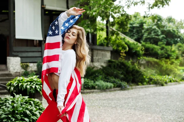 Una jovencita guapa se para en la calle y sostiene una gran bandera americana en sus manos —  Fotos de Stock