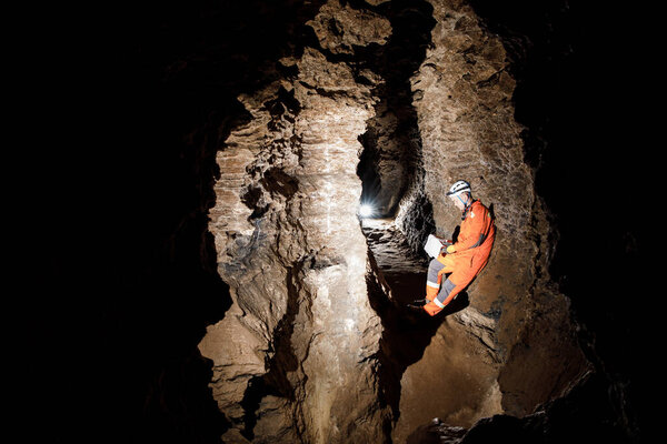 Man walking and exploring dark cave with light headlamp and map in his hand underground. Mysterious deep dark, explorer discovering mystery moody tunnel looking on rock wall inside.