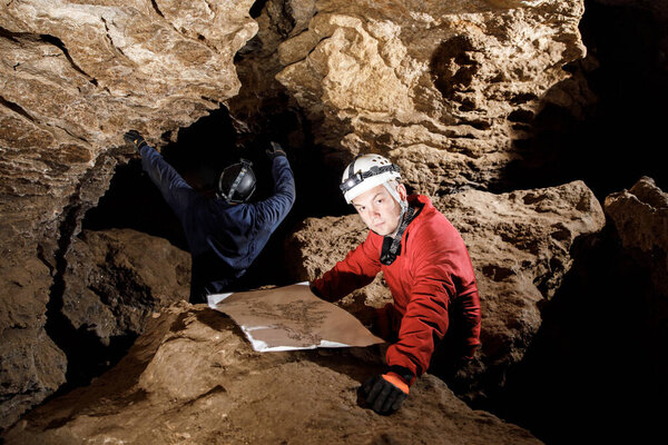 Two men, strong physique, explore the cave. Men dressed in special clothes to pass through the cave and stopped, looking at the map