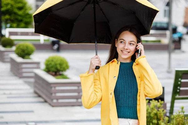Молода красива дівчина-підліток гуляє по місту в дощову погоду. Дівчина одягнена в кольоровий одяг і гумові черевики, що розмовляють по телефону — стокове фото
