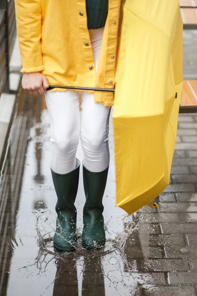 Дівчина, одягнена в кольоровий одяг і одягнена в гумові чоботи, стрибає в калюжу. Дівчина тримає жовту парасольку — стокове фото