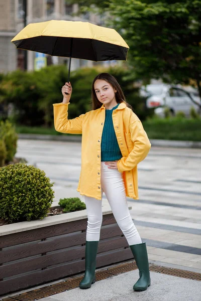 Молода красива дівчина-підліток гуляє по місту в дощову погоду. Дівчина одягнена в кольоровий одяг і гумові черевики — стокове фото