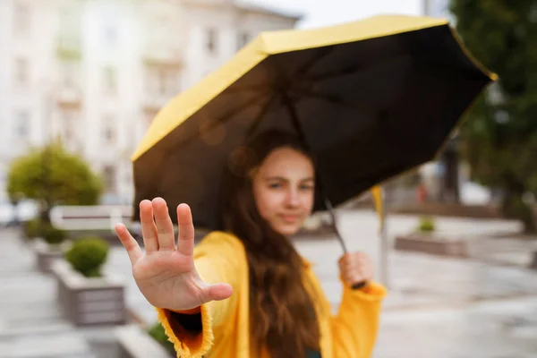 Молода красива дівчина-підліток гуляє по місту в дощову погоду. Дівчина, одягнена в кольоровий одяг і гумові чоботи, забороняє фотографу робити фотографії — стокове фото