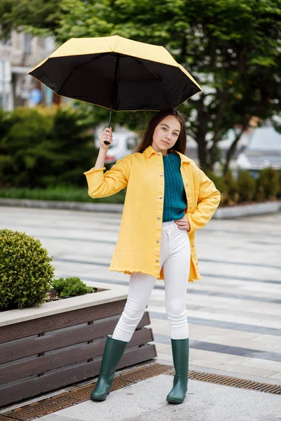 Молода красива дівчина-підліток гуляє по місту в дощову погоду. Дівчина одягнена в кольоровий одяг і гумові черевики — стокове фото