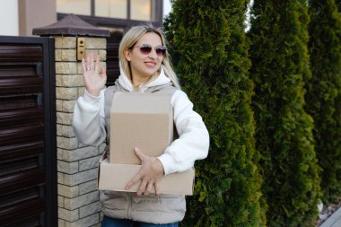 Elinde karton kutular ve evin girişinde güneş gözlüğü olan gülümseyen teslimatçı kadın.