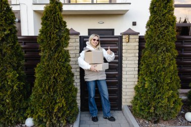 Karton kutular taşıyan teslimatçı kadın ve iskan girişinin önünde baş parmak işareti yapan kadın.