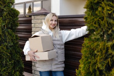 Elinde karton kutular ve evin girişinde güneş gözlüğü olan gülümseyen teslimatçı kadın.