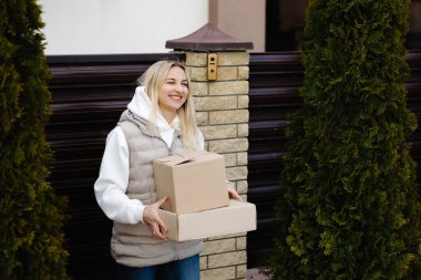 İki karton kutu taşıyan gülümseyen kadın evinin bahçesine giriyor ve iki thuja ağacının arasından geçiyor.