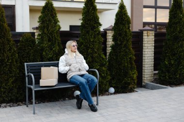 Genç bir kadın evinin dışındaki bankta oturuyor, paketlerle çevrili ve teslimat servisinin gelmesini sabırla bekliyor.