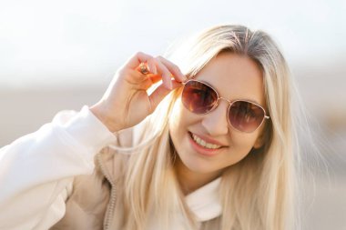 Güneşli bir günde şık güneş gözlüklerini indirirken parlak bir şekilde gülümseyen genç sarışın kadın mutluluk ve güzellik saçıyor.