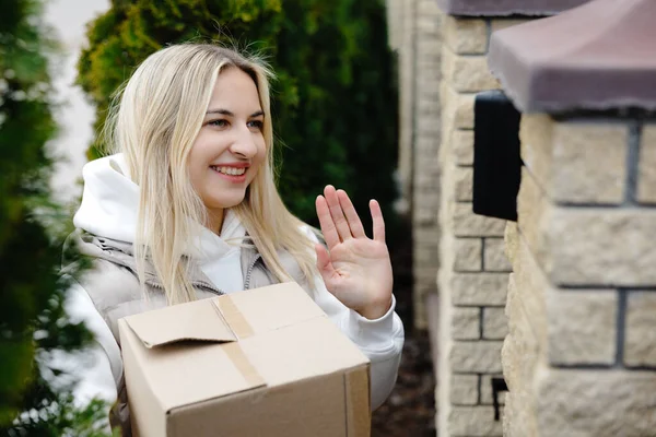 Teslimatçı kadın, elinde paketle evin girişinde gülümsüyor ve el sallıyor. Dışarıdaki dost canlısı müşteri hizmetlerini gösteriyor.