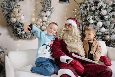 Noel Baba 'yla selfie çeken iki kardeş Noel ağacının yanındaki beyaz koltukta oturuyor.