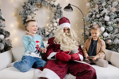 Noel Baba beyaz koltukta oturan iki süpriz çocuk ve onlara Noel ağacının yanında para veriyor.