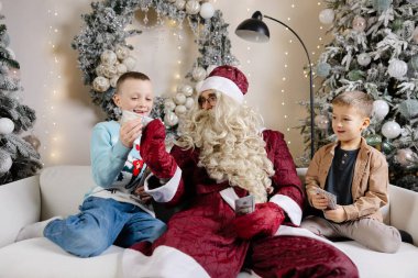 Noel Baba beyaz koltukta oturan iki süpriz çocuk ve onlara Noel ağacının yanında para veriyor.