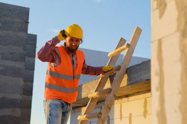 Güvenlik miğferi ve yeleği olan inşaat işçisi ahşap bir merdivene tırmanıyor, gün batımında inşaat çalışmalarını denetliyor.