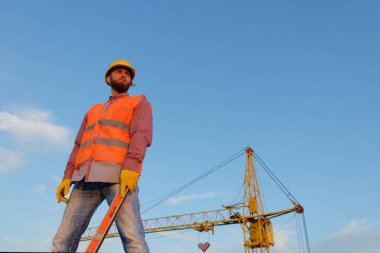 İnşaat işçisi, arka planda vinç bulunan inşaat alanında çelik yelek ve kask seviyesinde araç kullanıyor.