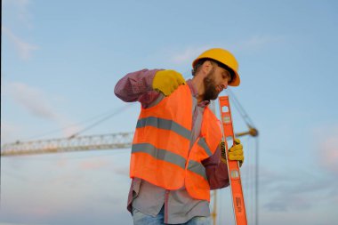 İnşaat işçisi yüksek görünürlük yeleği giyiyor ve arka planda vinç ile inşaat sahasında ruh seviyesini koruyor.