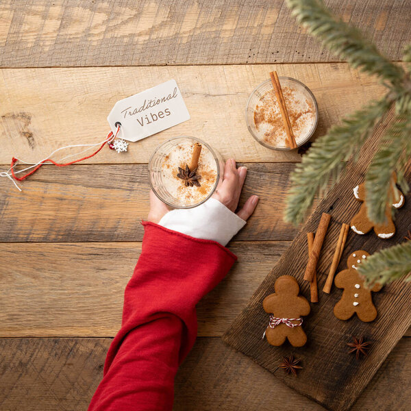 Hand reaching for a cup of Christmas holiday traditional eggnog on wooden table with gingerbread cookies, cinnamon sticks, star anise.  Message tag with text, "Traditional vibes."