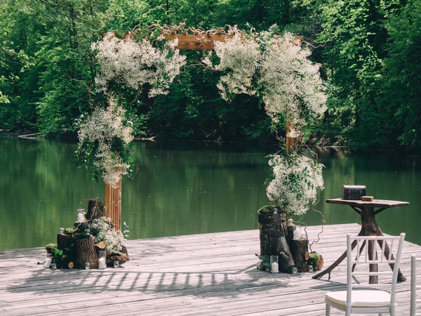 Romantic rustic wedding ceremony near river. White wooden chairs and flower arch.