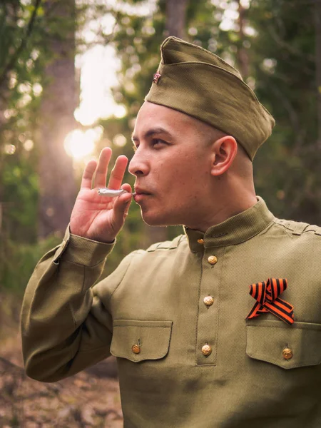 Man smokes in soviet war uniform in forest. Victory day reenactment