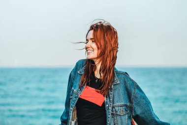 Kızıl saçlı güzel bir kız gülümsüyor. Modern gençlik. Rahat stil, kot giyim. Uzak mavi deniz. Kopya alanı. Güneş aydınlatması.